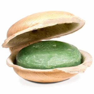 Un bar rotund, verde, de Săpun Holy Lama Yogi cu Vetiver în Frunză de Palmier, amplasat într-un recipient deschis, asemănător unei scoici, din material natural. Săpunul pare neted și translucid, infuzat cu aroma pământească a vetiverului, în timp ce recipientul are un aspect texturat, lemnos. Estetica generală este rustică și organică.