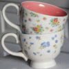 Două cești de ceai sunt stivuite una peste alta. Cupa de sus, o Cană din Ceramică Abby, 350 ml, are interiorul roz și este decorată cu flori colorate, în timp ce cea de jos are un design floral albastru. Fundalul are suprafețe din lemn alb, sugerând un cadru confortabil, familiar.