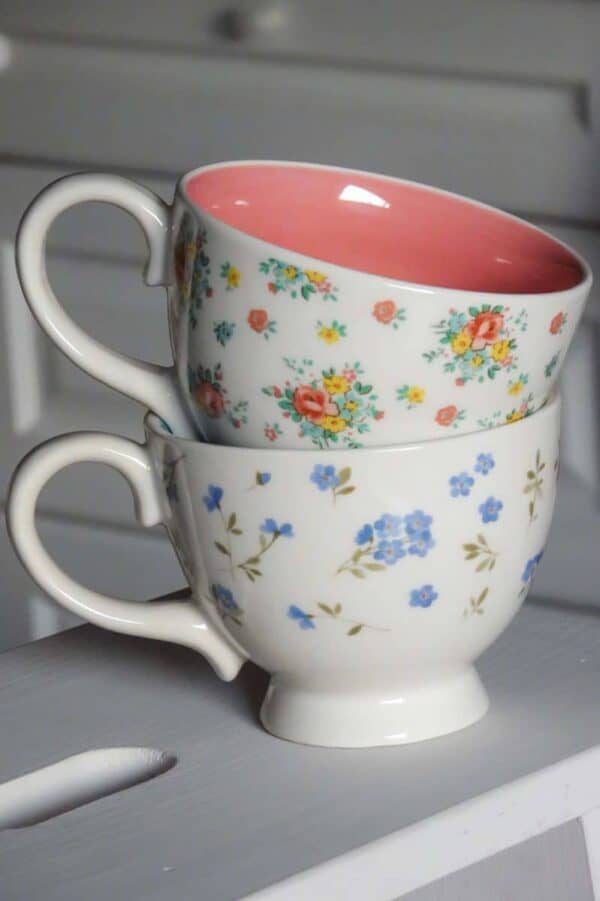 Două cești de ceai sunt stivuite una peste alta. Cupa de sus, o Cană din Ceramică Abby, 350 ml, are interiorul roz și este decorată cu flori colorate, în timp ce cea de jos are un design floral albastru. Fundalul are suprafețe din lemn alb, sugerând un cadru confortabil, familiar.