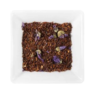 Un fel de mâncare pătrată albă conține frunze de ceai rooibos în vrac amestecate cu petale de flori violete uscate, care seamănă cu un amestec fin. Frunzele de ceai maro-roșcat și petalele violet creează un contrast atrăgător din punct de vedere vizual, perfect pentru asociere cu Brioșe cu Afine proaspăt coapte.