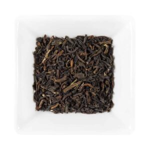 A white square dish fillled with loose-leaf Darjeeling a doua recoltare FTGFOP1 Amestec de Casă. Frunzele uscate de ceai sunt maro închis, cu un aspect ușor răsucite, ceea ce indică că sunt frunze pline ale recoltei renumite. Fundalul este alb simplu, subliniind textura și culoarea frunzelor de ceai.